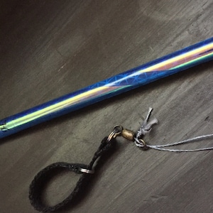 Può includere: Una bacchetta blu e iridescente con un cinturino nero e una fibbia argentata. La bacchetta è attaccata a uno spago bianco.