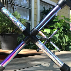 Può includere: Uno sgabello pieghevole a forma di X con gambe iridescenti color arcobaleno. Le gambe sono collegate da giunti in plastica nera e hanno piedini in gomma neri. Le gambe hanno un motivo geometrico argentato.