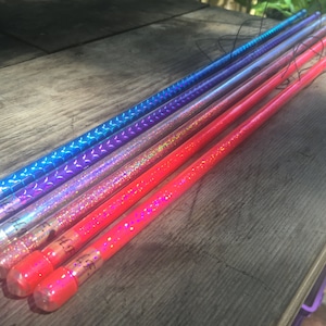 Può includere: Una collezione di sette bacchette colorate, ognuna con un design unico. Le bacchette sono in tonalità di blu, viola e rosso, con dettagli glitterati e fantasia. Sono disposte su una superficie di legno.