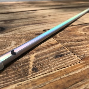 Può includere: Una bacchetta argentata iridescente con un design a forma di cuore nero. La bacchetta è appoggiata su una superficie di legno.