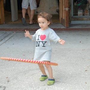 May include: A young person wearing a grey shirt with the words "I NY" and a red heart printed on it, is playing with a pink and yellow hula hoop. They are wearing green shoes.