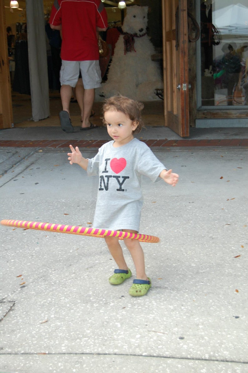 May include: A young person wearing a grey shirt with the words "I NY" and a red heart printed on it, is playing with a pink and yellow hula hoop. They are wearing green shoes.