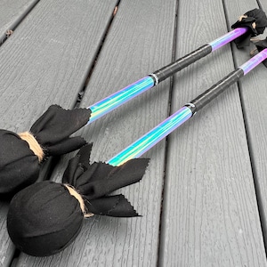 Può includere: Due oggetti di scena per lo spinning poi. Ogni poi ha una testa in tessuto nero legata con spago, un'impugnatura avvolta in nero e un'asta olografica iridescente. Gli oggetti di scena sono appoggiati su una superficie di legno grigio.