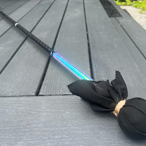 Puede incluir: Un bastón de fuego negro con una sección iridiscente y una bola de tela negra. El bastón tiene un agarre texturizado y un acento rosa. La bola está atada con hilo. El bastón mide aproximadamente 100 cm de largo.