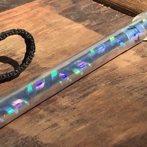 Może przedstawiać: Przezroczysta, cylindryczna r&oacute;żdżka z iryzującymi, kolorowymi akcentami. R&oacute;żdżka ma przezroczystą, zaokrągloną końc&oacute;wkę i czarną pętlę ze sznurka. R&oacute;żdżka leży na drewnianej powierzchni.