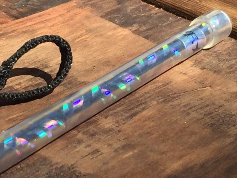 Może przedstawiać: Przezroczysta, cylindryczna r&oacute;żdżka z iryzującymi, kolorowymi akcentami. R&oacute;żdżka ma przezroczystą, zaokrągloną końc&oacute;wkę i czarną pętlę ze sznurka. R&oacute;żdżka leży na drewnianej powierzchni.