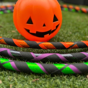 Può includere: Tre cerchi da hula colorati con nastro nero, arancione, verde e viola. I cerchi sono sdraiati sull'erba verde con una zucca arancione sullo sfondo.