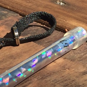 Może przedstawiać: Przezroczysta, cylindryczna tuba z iryzującymi, geometrycznymi kształtami w środku i przezroczystą nasadką. Na tubie znajduje się napis "FF". Obok tuby znajduje się czarna, tkana pętla z metalowym pierścieniem.