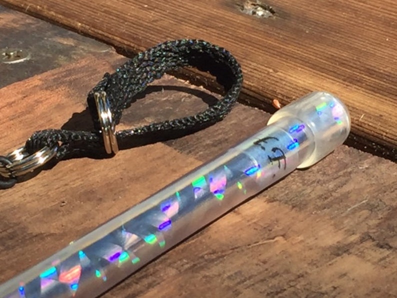 Może przedstawiać: Przezroczysta, cylindryczna tuba z iryzującymi, geometrycznymi kształtami w środku i przezroczystą nasadką. Na tubie znajduje się napis "FF". Obok tuby znajduje się czarna, tkana pętla z metalowym pierścieniem.