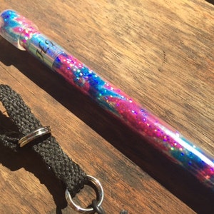 Puede incluir: Una varita de brillo rosa y azul con un tubo de plástico transparente y una tapa plateada. La varita tiene una correa negra con un anillo plateado.