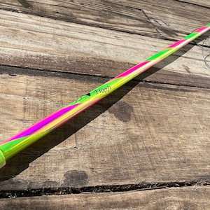 Può includere: Un bastone lungo, sottile, verde, rosa e giallo con un logo nero che dice "WOOD". Il bastone ha un tappo verde a un'estremità.