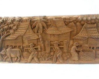 May include: A hand-carved wooden wall art depicting a traditional Thai village scene. The artwork features intricately carved houses, palm trees, and figures of people going about their daily lives.