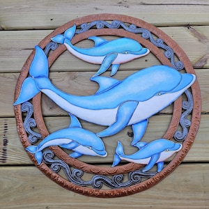 May include: A wooden wall art piece featuring a circular design with four blue dolphins swimming within a brown frame. The dolphins have white bellies and are set against a wooden background.