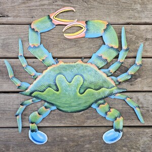 May include: A metal wall art piece shaped like a crab, featuring a gradient of blue, green, and yellow hues. The crab has two large pincers at the top, eight legs extending from the sides, and a textured shell with a distinct central ridge.