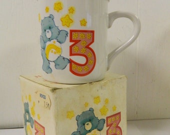 May include: A white ceramic mug with a blue Care Bear holding a rainbow and a pink number 3. The mug is sitting on a white box with a similar design. The box has the word "REYCO" printed on it.