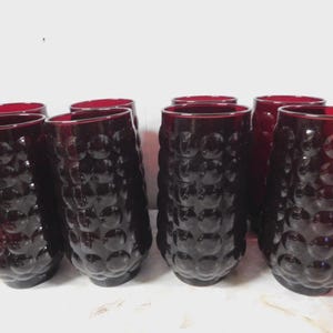 May include: Set of eight ruby red drinking glasses. Each glass has a textured, bubble-like pattern on the exterior. The glasses are cylindrical with a slightly flared rim. The glasses are arranged on a white surface.