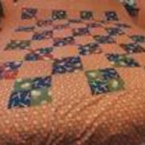 May include: A quilt featuring a repeating pattern of orange fabric squares with small white dots, interspersed with dark blue and green squares that display a repeating pattern of white star shapes and various small, colorful, indistinct graphic icons.