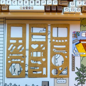 Puede incluir: Tres plantillas de papel marrón con varias formas recortadas, incluyendo rectángulos, corazones y círculos. Un teclado y una alfombrilla decorativa con una ilustración de dibujos animados están en el fondo. También es visible una pequeña pegatina con el texto "BEAUTYBYTABLET".