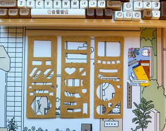 May include: Three brown paper stencils with various cut-out shapes, including rectangles, hearts, and circles. A keyboard and a decorative mat with a cartoon illustration are in the background. A small sticker with the text "BEAUTYBYTABLET" is also visible.