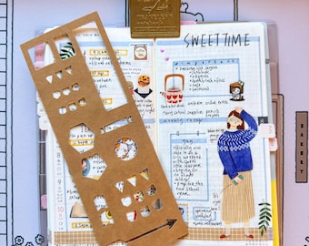 May include: A planner with the text "SWEET TIME" and a brown paper stencil. The planner has a gold metal clip and is open to a page with handwritten notes and illustrations. The background features a keyboard and a patterned mat.