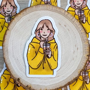 May include: A sticker featuring an illustration of a person in a yellow hoodie holding a bubble tea. The person has brown hair and is wearing pink earrings. The stickers are arranged on a wooden slice.