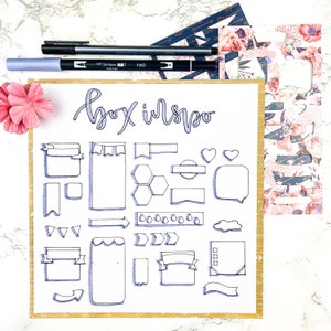 May include: A white paper with the handwritten text "box inspo" and various hand-drawn shapes, banners, and icons in blue ink. Two pens and decorative paper are on top. A pink flower is in the corner.