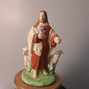 May include: A porcelain figurine of Jesus holding a lamb, with two other lambs at his feet. The figurine is on a wooden music box base with the inscription "The Lord is my Shepherd".