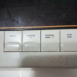 May include: Close-up of a white control panel with four rectangular buttons. The buttons are labeled "ANSWER", "MESSAGE", "PERSONAL MEMO", and "STOP/SAVE". The panel has a black top section and an orange stripe.