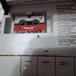 May include: A vintage white answering machine with a red, white, and black Panasonic RT-60MC microcassette tape. The machine has buttons labeled "Answer", "Message", and "Personal Memo". Brief operating instructions are visible.