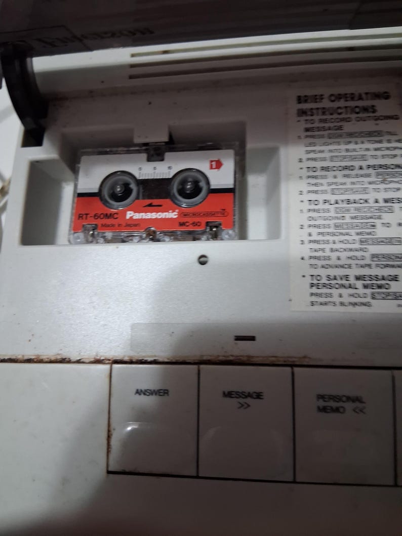 May include: A vintage white answering machine with a red, white, and black Panasonic RT-60MC microcassette tape. The machine has buttons labeled "Answer", "Message", and "Personal Memo". Brief operating instructions are visible.