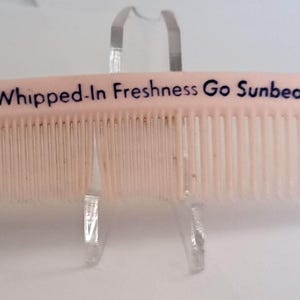 May include: A pale pink comb with closely spaced teeth, bearing the text "For Whipped-In Freshness Go Sunbeam Bread" in black. The comb is presented on a clear stand, highlighting the design and the brand's message.