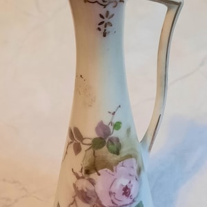 May include: A white porcelain jug with a delicate floral design featuring pink roses and green leaves. The jug has a gold trim around the handle and a gold design around the neck. The jug is signed "P.K. & Co."
