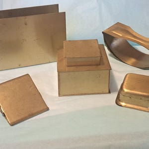 May include: A set of six gold-colored metal boxes and a metal ring. The boxes are different sizes and shapes, and some are stacked on top of each other. The ring is curved and has a smooth surface.