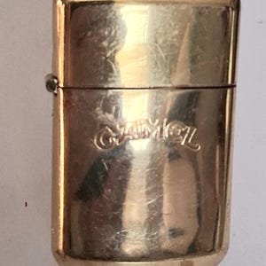 May include: A vintage gold-colored metal lighter with the word "CAMEL" embossed on the front. The lighter has a rectangular shape with rounded edges and a hinged lid. The lighter shows signs of wear.