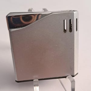 May include: Silver lighter with a rectangular shape and a smooth, polished finish. It has three small, rectangular holes on the side.