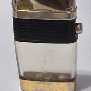 May include: A vintage rectangular lighter. It features a gold-coloured top and bottom, a clear central section, and a black rubber band around the middle. The lighter has a small, round button on the side.