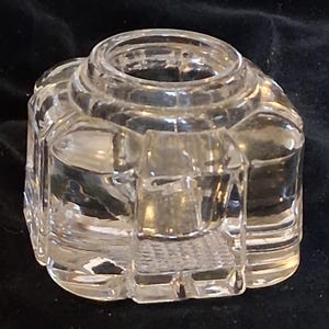 Puede incluir: Un tintero de vidrio transparente y facetado con una abertura cilíndrica y un diseño geométrico. El tintero tiene una base cuadrada con esquinas redondeadas y un patrón texturizado en la parte inferior.