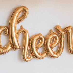 May include: Gold foil letter balloons spelling out the word "cheers".
