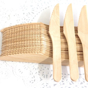 May include: A stack of disposable wooden knives, with three knives standing upright in front of the stack. The knives are made of natural wood and have a serrated edge.