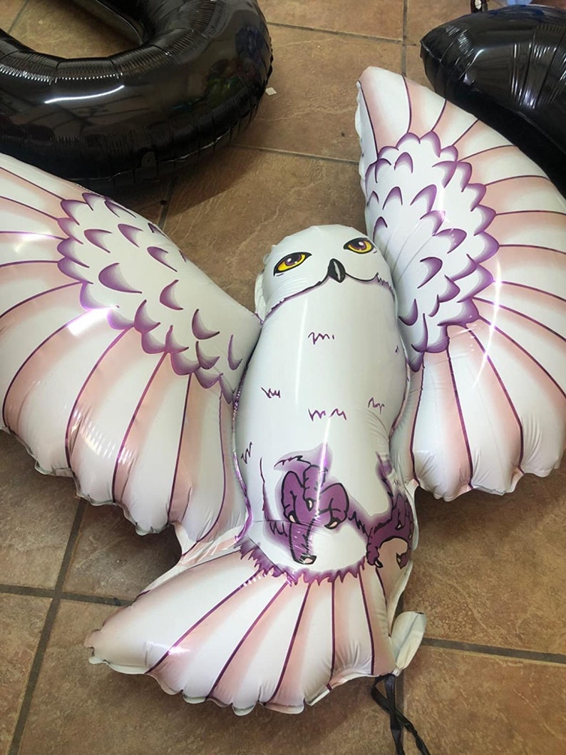May include: A large, white owl-shaped balloon with outstretched wings. The balloon features pink and purple accents on the wings and a cartoon-style owl face with yellow eyes. Black balloons are visible in the background.