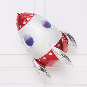 May include: A red and silver foil rocket balloon with white polka dots and blue and purple windows. The balloon is inflated and ready to be used for a party or celebration.