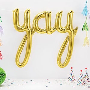 May include: Gold foil letter balloons spelling "yay" hanging from a white string with colorful tassels on either side.