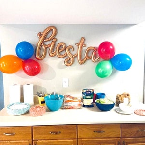 May include: A kitchen counter with a variety of food and party supplies. There are bowls of food, a bag of cheese, paper towels, and a gold foil "fiesta" banner with colorful balloons.