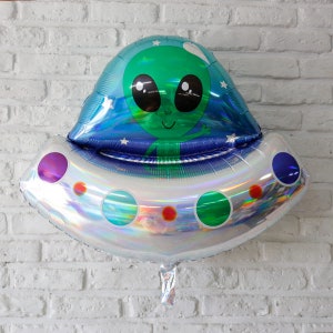May include: A silver holographic foil balloon shaped like a UFO with a green alien inside. The alien has big eyes and a friendly smile. The UFO has purple and blue accents and a string for hanging.
