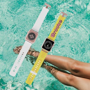 May include: Two Apple Watch bands, one white and one yellow with a pink "Brooke" design. The yellow band has a pineapple pattern.
