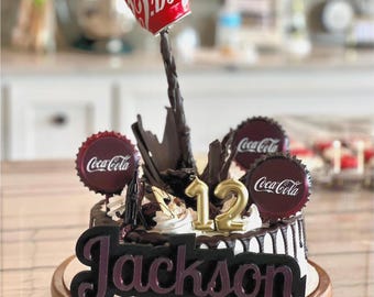 May include: A round cake decorated with a red Coca-Cola can tilted to pour a dark liquid stream, two circular Coca-Cola bottle cap toppers, a gold number 12 candle, and a black sign with the name Jackson in a script font on the front.