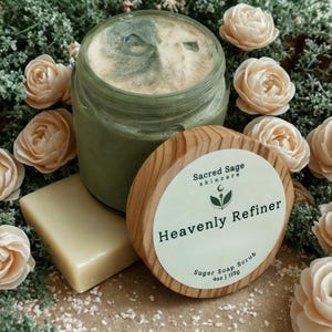 May include: A jar of green sugar soap scrub with a wooden lid, a bar of soap, and decorative flowers. The lid reads "Heavenly Refiner" and "Sugar Soap Scrub 4oz | 113g". The jar is surrounded by cream-colored flowers and greenery.