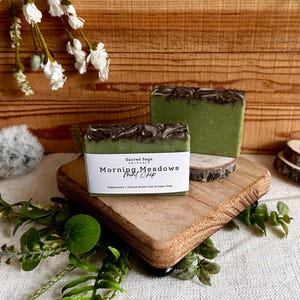 May include: Two green artisan soaps with a dark brown swirl design on top, labeled "Morning Meadows Mint Chip." The soaps sit on a wooden board and a small wooden slice. The soaps are surrounded by greenery and white flowers.