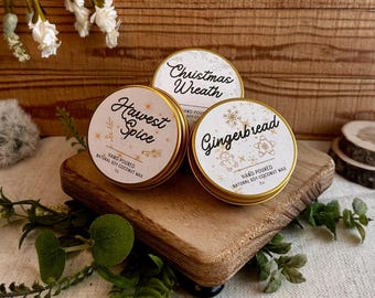May include: Three round, gold-rimmed tins of hand-poured soy coconut wax candles. The labels read "Harvest Spice", "Christmas Wreath", and "Gingerbread". The tins sit on a small wooden stand with greenery and white flowers in the background.