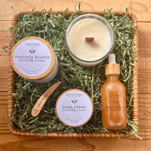 May include: A gift basket with skincare products, including a jar of Heavenly Soufflé sugar soap scrub, a candle, a bottle of Shiloh Oasis oil, and Selah Crème skin moisturizer. The products are arranged on green paper in a woven basket.